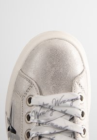 Metallisk silver sneaker med en strukturerad yta, vit gummisula och tryckta snören med grå handskriven stil.