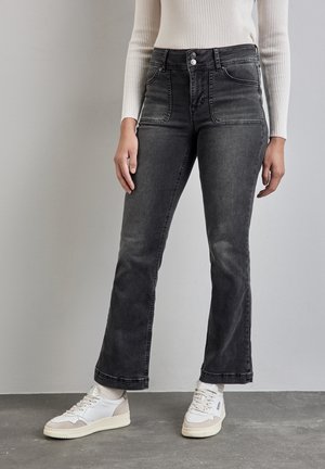 Femme portant un jean noir taille haute avec poches avant, un top crème côtelé à manches longues, des baskets blanches et des chaussettes blanches, debout sur un sol gris.