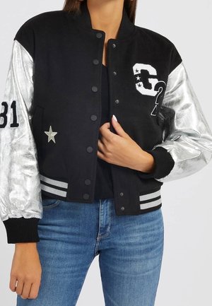 Vrouw draagt een zwart-zilveren varsityjack met mouwnummer 31, letter- en sterapplicaties, over een zwarte top en blauwe jeans.