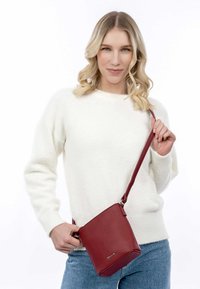 Bolso cruzado de cuero rojo con un acabado texturizado y correa larga, sostenido por una persona con un suéter blanco. Diseño simple y cuadrado con acentos de metal.
