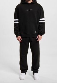 Zwarte hoodie met witte strepen op de mouwen, een voorzak en een geborduurd logo. Gepaard met zwarte sweatpants en witte sneakers.