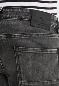 Unos vaqueros de mezclilla negra con un aspecto desgastado, que presentan una etiqueta de cuero que dice "SCOTCH&SODA AMSTERDAM'S BLAUW" y dos bolsillos traseros.