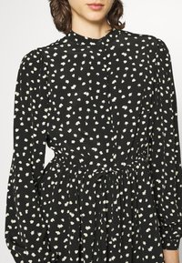 Vestido negro con un pequeño patrón floral blanco, mangas largas, cuello alto y cintura con cordón para un ajuste ajustable. Textura de tela suave.