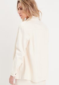 Veste à boutons de couleur crème clair, en tissu doux. Présente une coupe décontractée, des manches longues et deux poches avant avec des détails de coutures subtils.