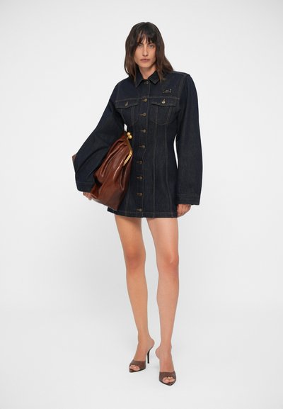 Πουκαμίσα φορέματος από τζιν σε σκούρο μπλε χρώμα με κουμπιά μπροστά, μυτερό γιακά και δύο τσέπες στο στήθος. Συνδυάζεται με καφέ δερμάτινη τσάντα και ψηλοτάκουνα παπούτσια.