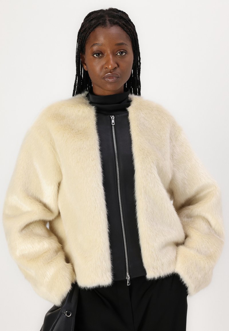 Kremowa kurtka z sztucznego futra z szerokim kołnierzem, miękką teksturą i luźnym krojem. Noszona na czarnej bluzie zapinanej na zamek, z długimi rękawami i minimalistycznym designem.