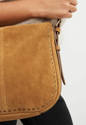 Bolso bandolera de ante color canela con tachuelas de latón a lo largo del borde, sostenido por una mano que lleva un suéter crema y pantalones negros.