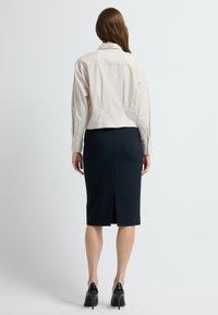 Kobieta stojąca tyłem, ubrana w beżową koszulę z długim rękawem, czarną spódnicę o długości do kolan oraz czarne szpilki na jednolitym tle.