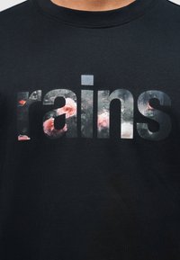 Μαύρο πουκάμισο με τη λέξη «rains» σε μεγάλα γράμματα γεμάτα με ένα λουλουδάτο μοτίβο που περιλαμβάνει ροζ τριαντάφυλλα και πράσινα φύλλα.