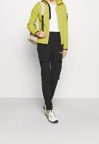 Grüne Zip-Jacke, weißes T-Shirt, schwarze Hose mit Seitentaschen und weiße Sneakers mit neon-gelben Akzenten. Neutraler Hintergrund.