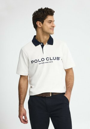 Uomo che indossa una maglia Polo Club bianca con colletto blu navy e pantaloni blu navy, in piedi con una mano in tasca, che guarda verso sinistra.