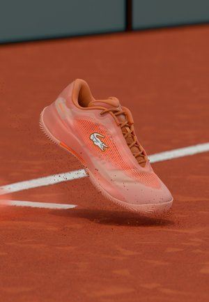 Zapatilla de tenis naranja con parte superior de malla, que presenta un logo blanco, suela texturizada y diseño dinámico, mostrada sobre una superficie de pista de tierra batida.