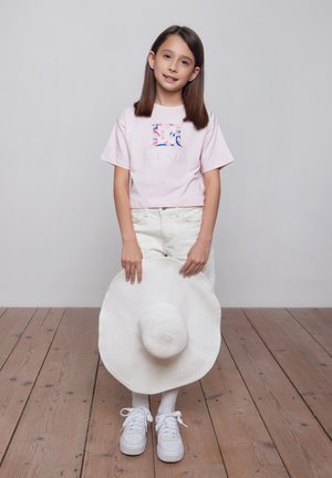 Giovane ragazza con capelli castani lisci che indossa una maglietta rosa e pantaloni bianchi, tenendo davanti a sé un grande cappello bianco da sole.