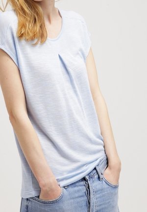 Kobieta w jasnoniebieskiej koszuli z krótkim rękawem z plisą z przodu i jasnych dżinsach, ręce w kieszeniach, na tle jasnego, gładkiego tła.