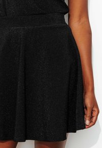 Jupe évasée noire en tissu texturé avec un léger éclat. La ceinture est élastique et l'ourlet arrive à mi-cuisse.