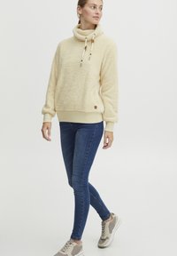 Cream-colored, fuzzy fleece pullover with a cowl neck, drawstring closure, ribbed cuffs, and a small logo patch on the hem. Paired with blue jeans.