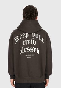 Person, der einen dunklen Kapuzenpulli mit dem Schriftzug "Halt deine Crew gesegnet" und kleinerer Aufschrift über Vertrauen auf dem Rücken trägt, steht vor einem schlichten Hintergrund.