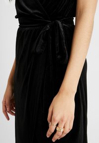 Schwarzes Samtkleid mit einer gebundenen Taillendetail. Nahaufnahme zeigt eine Hand mit mehreren goldenen Ringen an schlanken Fingern. Die weiche Textur wird hervorgehoben.