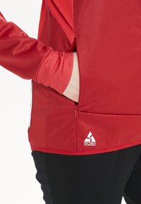Röd sportjacka med texturerad yta, sidoficka och WX-TECH-logotyp. Har långa ärmar och en dragkedjeöppning vid nederkanten.