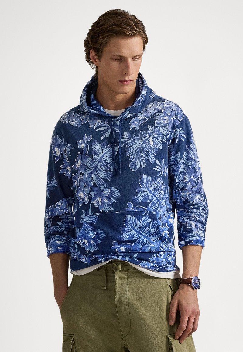 Homem usando hoodie azul com estampa floral, calças cargo verde-oliva e relógio no pulso, olhando para baixo contra fundo liso.