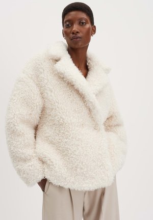 Giacca bianca, soffice e strutturata con un grande colletto e tasche frontali, indossata su pantaloni leggermente beige. Materiale morbido e accogliente.