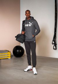 Sudadera con capucha gris y logo blanco, pantalones oscuros a juego y zapatillas blancas con acentos rojos y azules, sosteniendo un bolso de deporte negro.