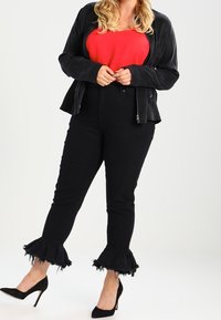 Débardeur rouge, veste noire zippée, jeans noirs avec des bords effilochés et évasés, et talons hauts noirs. Texture lisse, design ajusté.