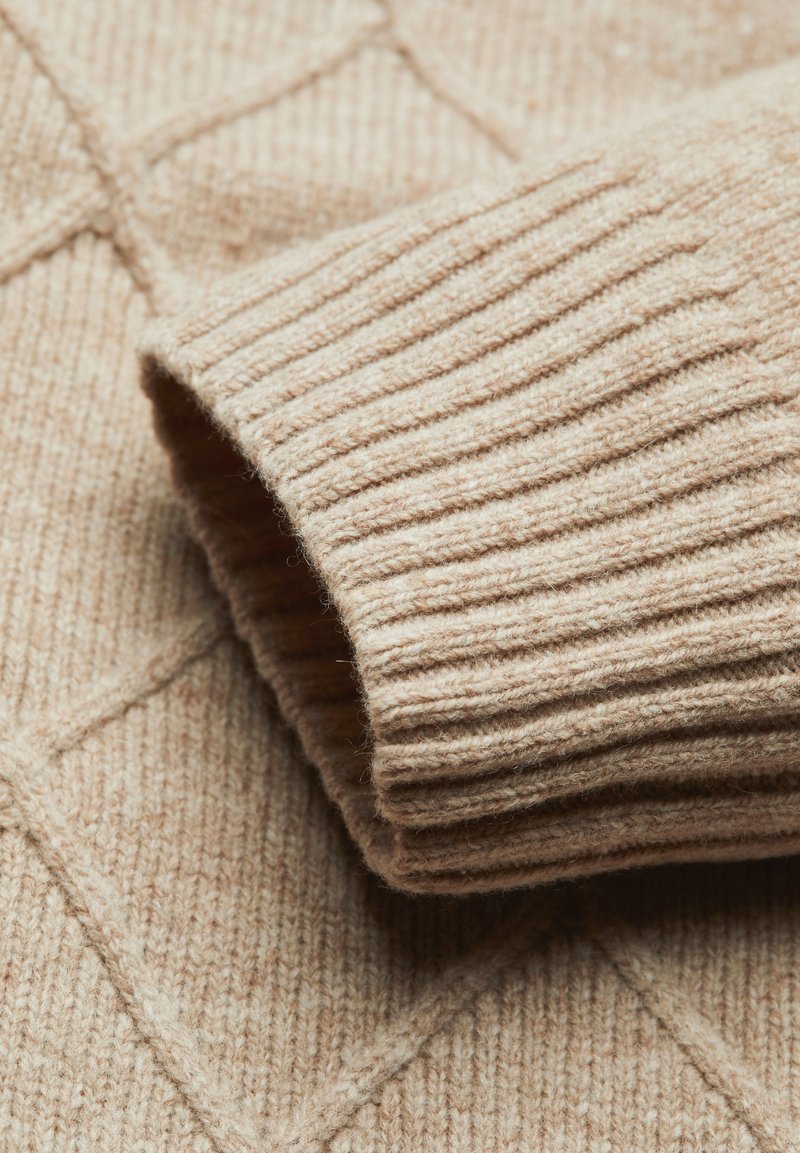 Maglione a collo alto beige a maglia con motivo a diamante, caratterizzato da polsini a coste e una consistenza morbida. Primo piano della manica contro il corpo del capo.