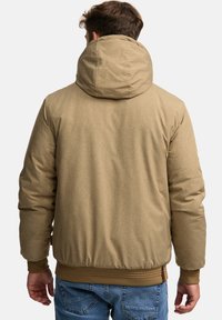 Pufferjakke i lysbrunt stof med en tekstureret overflade, udstyret med hætte og elastiske manchetter, parret med blå jeans.