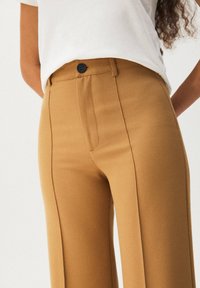 Brązowe, dopasowane spodnie wykonane z gładkiej tkaniny, z przednim suwakiem i pojedynczym zapięciem na guzik. Projekt zawiera boczne kieszenie.