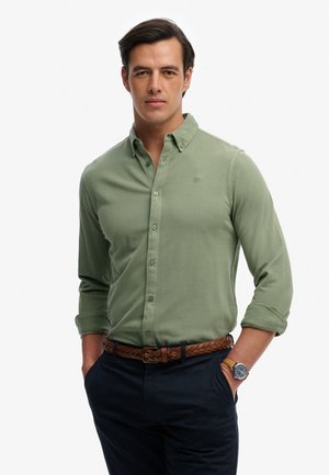 Man draagt een lichtgroen overhemd met opgerolde mouwen, donkere broek, gevlochten bruine riem en een zandkleurig horloge, staat met handen in de zakken.