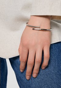 Srebrna metalowa bransoletka na nadgarstek o eleganckim, minimalistycznym wzornictwie. Posiada gładką, polerowaną powierzchnię i asymetryczne zapięcie.