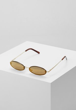 Occhiali da sole ovali marroni con lenti colorate, sottili montature in metallo dorato e punte laterali in plastica marrone, appoggiati su una superficie bianca.