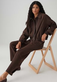 Bruine jas met rits aan de voorkant, cropped ontwerp, elastische tailleband; bijpassende bruine broek met een losse pasvorm en omgeslagen zoom, gedragen met instapschoenen.