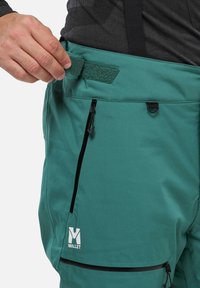 Türkise Outdoor-Hosen mit Velcro-Verstellung an der Taille, seitlichen Reißverschlusstaschen und einem weißen Logo unten links. Glatte Textur.