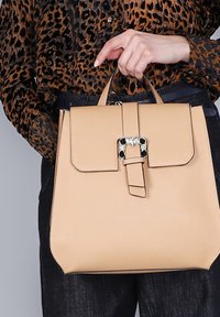 Mochila de cuero en color beige con una forma estructurada. Cuenta con un cierre de solapa, asas ajustables y una hebilla decorativa con piedras preciosas.