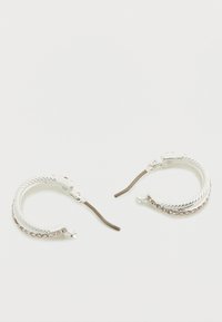 Ασημένια σκουλαρίκια κρίκοι με σχεδιασμό από πλεγμένο σχοινί. Διαθέτουν υφή στην επιφάνεια και διακοσμητική αλυσίδα κατά μήκος της άκρης. Κλείσιμο με ασφαλές κούμπωμα.