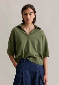 Zielony, oversize'owy sweter z dekoltem w serek i krótkimi rękawami, połączony z ciemnoniebieską spódnicą z wysokim stanem z denimu, z plisami.