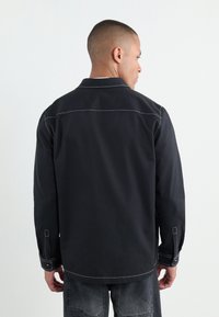 Schwarzes Langarmhemd mit kontrastierenden weißen Nähten, geknöpften Manschetten und klassischem Kragen. Der Stoff erscheint glatt und leicht.