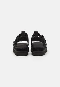 Zwarte sandalen met een open achterkant, voorzien van textuurbanden, verstelbare gespen en een platte rubberen zool. UGG-logo zichtbaar op de hiel.