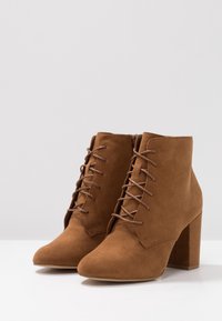 Bruna mocka ankelskor med spetsig tå, snörning framtill och klack i blockformat. Har en slät yta och en enkel design.
