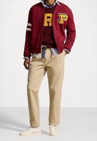 Burgundy kofta med gula och vita detaljer, här över en vinröd tröja. Beiga byxor och vita sneakers fulländar outfiten.