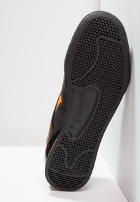 Černá podrážka tenisky s texturovaným, tečkovaným gripem. Obsahuje značku Reebok, s oranžovými akcenty a hladkým okolním svrškem.