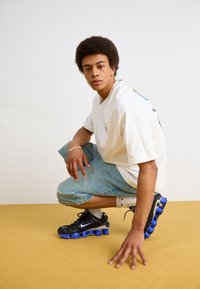 Zapatillas deportivas negras con acentos azules, combinadas con pantalones cortos de mezclilla azul claro y una camiseta blanca holgada, sobre un fondo amarillo.