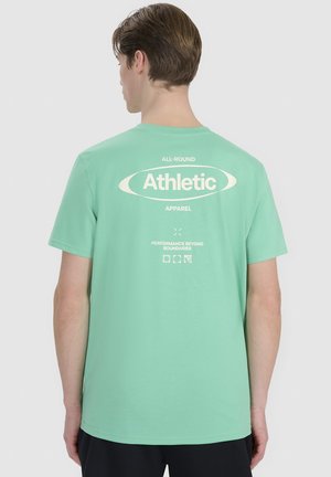 Młody mężczyzna z krótkimi brązowymi włosami, ubrany w miętowy T-shirt z białym napisem marki Athletic na plecach, stojący na jednolitym tle.