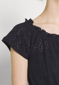 Svart bomullblus med korta volangärmar, med ögonletsperforeringar, elastisk halsringning och en mjuk textur.