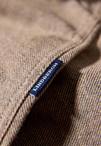 Gestructureerde bruine stof met subtiel visgraatpatroon, voorzien van een blauwe label met het woord "LINDBERGH" in het wit.
