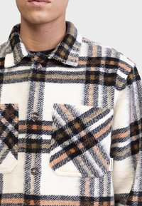 Chemise en flanelle à carreaux dans les tons crème, noir, orange et gris. Présente un col, une fermeture à boutons et deux poches poitrine avec une finition texturée.