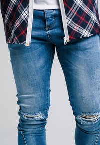 Primer plano de una persona que lleva jeans azules desgastados y una chaqueta de cuadros en rojo, negro y blanco sobre una camiseta blanca.