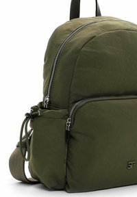 Mochila verde oliva con una tela suave y texturizada, con cremalleras plateadas, múltiples compartimentos y una correa ajustable para mayor comodidad.